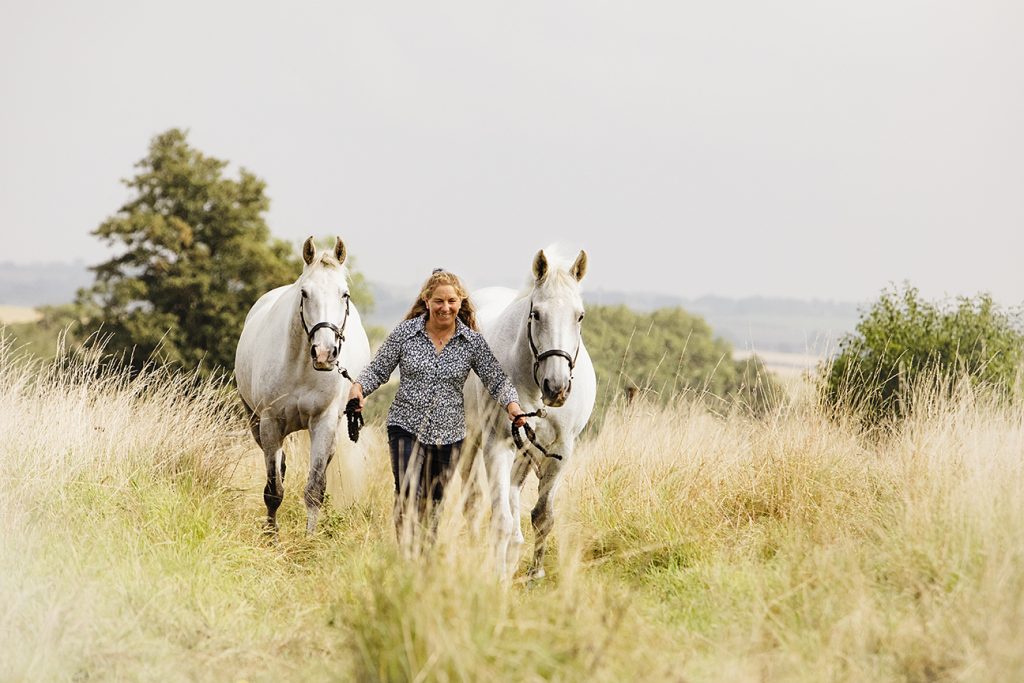 Equestrian leads her two grey horses across a Northamptonshire field of long grass