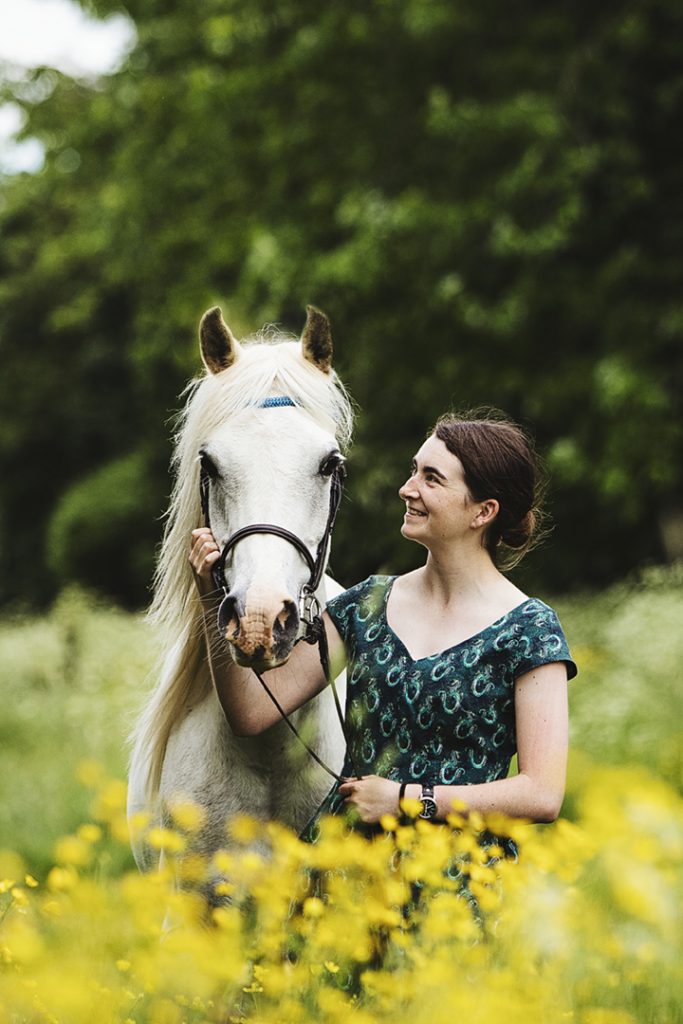 Rider wearing a green dress stands in a field of yellow flowers with her grey pony